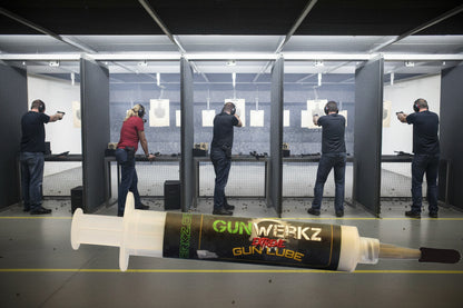 1/3 ounce syringe of Gun Werkz Extreme Gun Lube with an indoor range with 1 woman and 4 men taking aim at the targets as the background
